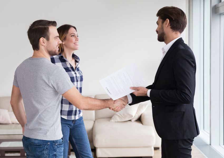 A property manager in a black suit holds paperwork while shaking hands with two new tenants during tenant screening.